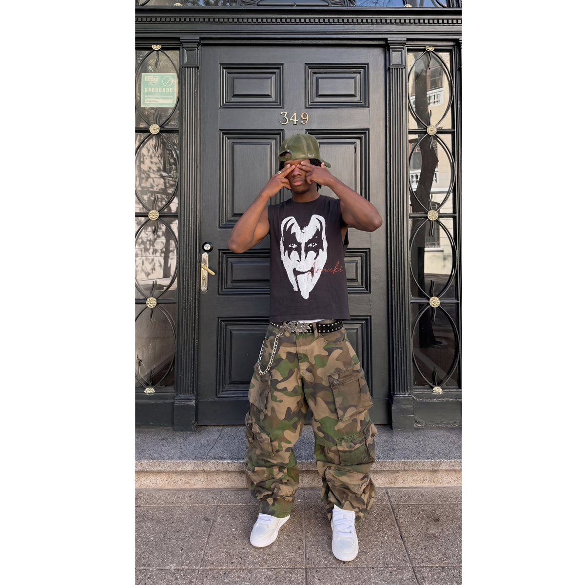Person wearing a black t-shirt with a white logo and camouflage pants standing in front of a black door.